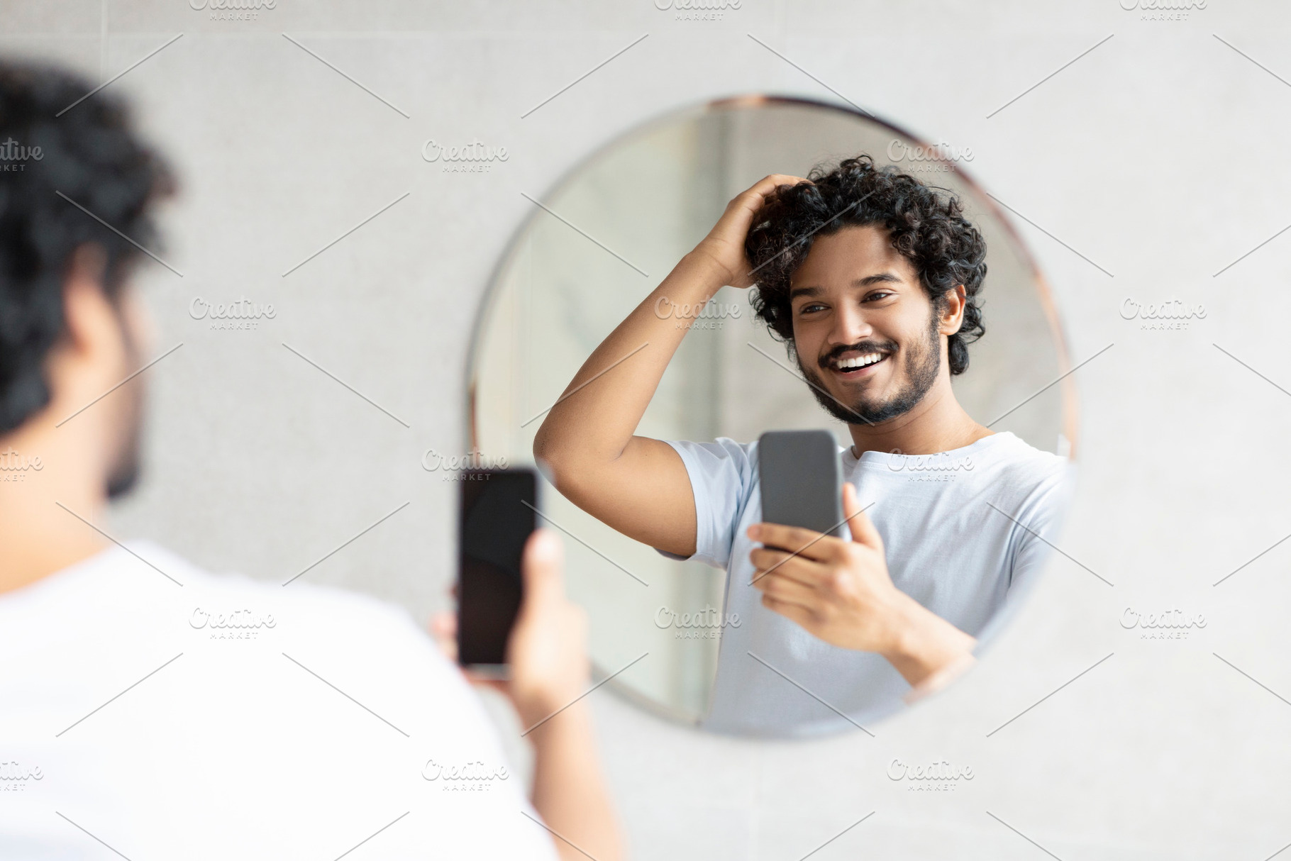 Cheerful indian man standing next to mirror in modern bathroom, holding smartph, a Person Photo by Prostock-Studio