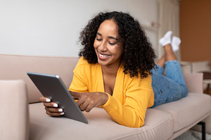 Relaxed young black woman lying on couch and using tablet pc, chatting online o, a Photo by Prostock-Studio