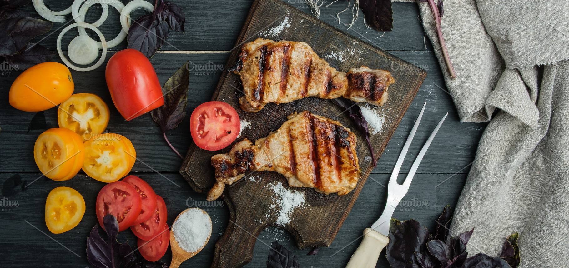 Grilled pork steaks on a wooden board, surrounded by vegetables, a Food & Drink Photo by ndanko