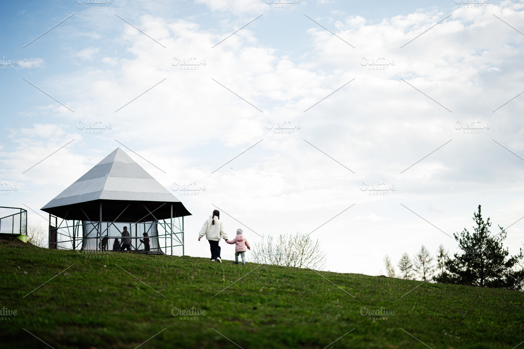 Girl and child walking hand in hand toward a pavilion on a grassy hillside in a, a Person Photo by AS photostudio