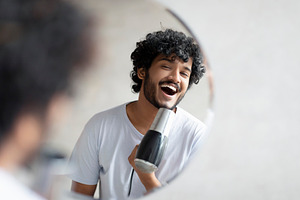 Joyful indian man holding hairdryer and singing as in microphone, having fun wh, a Person Photo by Prostock-Studio