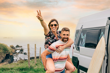 Couple looking camera having fun piggybacking next to their camper van, a Holiday Photo by davidpereiras