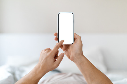 Male hands holding phone with white empty screen, bedroom interior, a Photo by Prostock-Studio