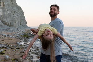 Smiling man holding young girl upside down on a rocky beach, happy family momen, a Person Photo by Ales Munt