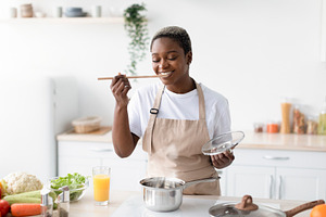 Smiling millennial african american woman in apron cooking at home and tasting, a Person Photo by Prostock-Studio