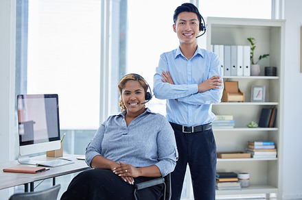 Portrait of two happy smiling diverse call centre telemarketing agents wearing headsets while working in an office. Confident and friendly african american and asian assistants operating helpdesk for customer service support, a Photo by Peopleimages.com