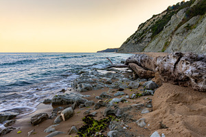 Serene rocky shoreline with gentle waves, driftwood, and a dramatic cliff at su, a Background Photo by Ales Munt