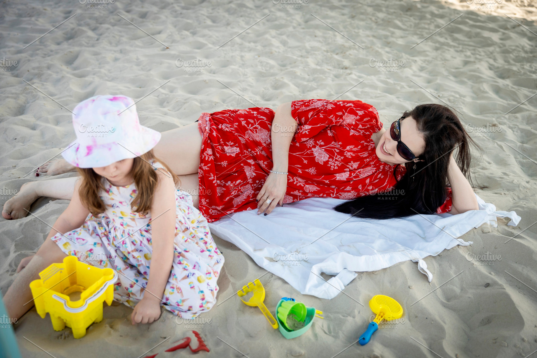 Mother relaxing on a towel on the beach while her daughter plays, a Person Photo by Imaginarium
