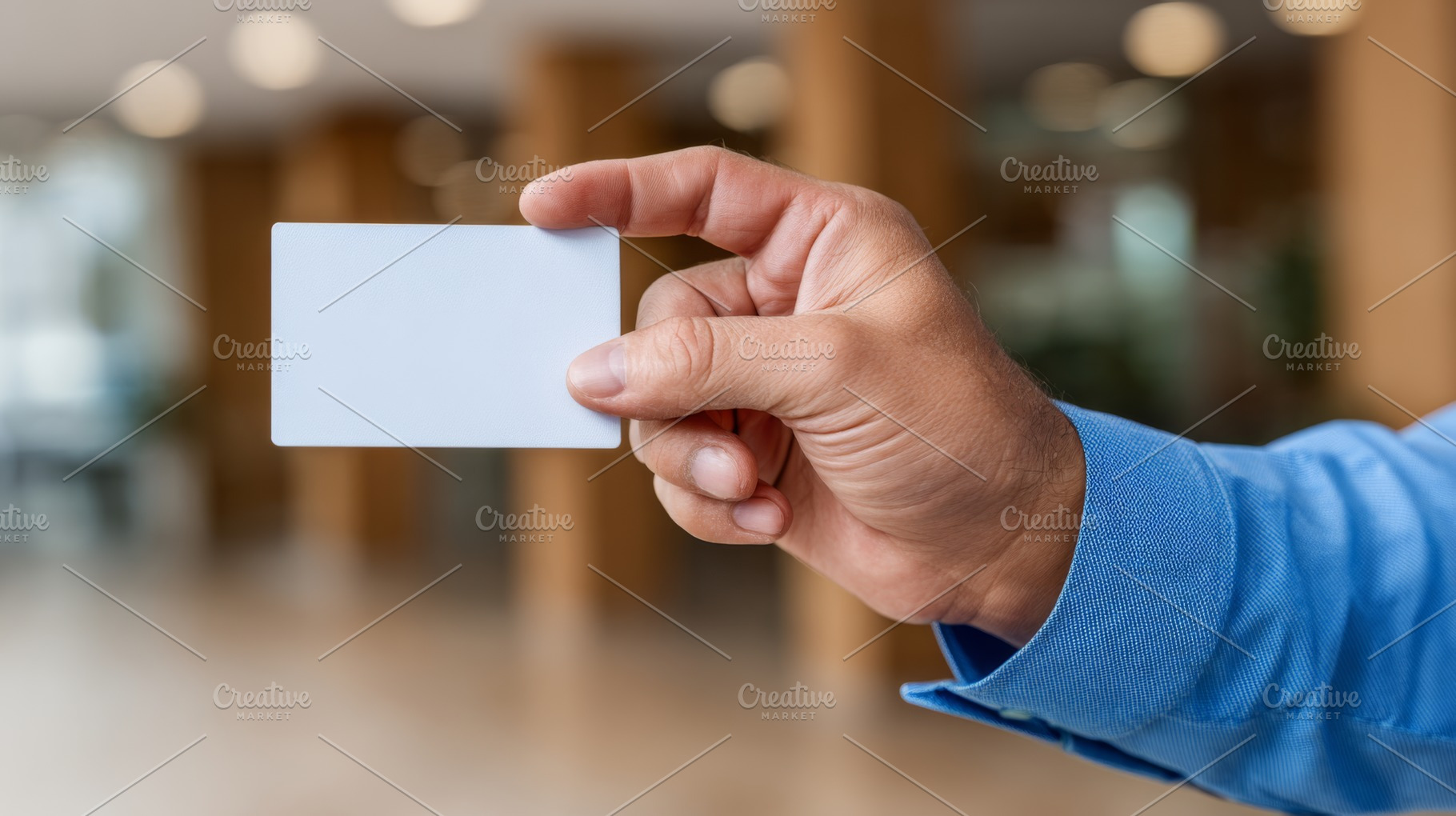 A male hand holding a blank business card, set against a blurred interior backg, a Business Photo by ImageMir