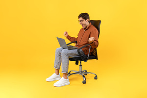 Joyful Guy Shaking Fists Using Laptop Sitting Over Yellow Background, a Photo by Prostock-Studio
