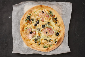 Round pizza with bacon, chicken, olives and cheese on parchment, a Food & Drink Photo by ndanko