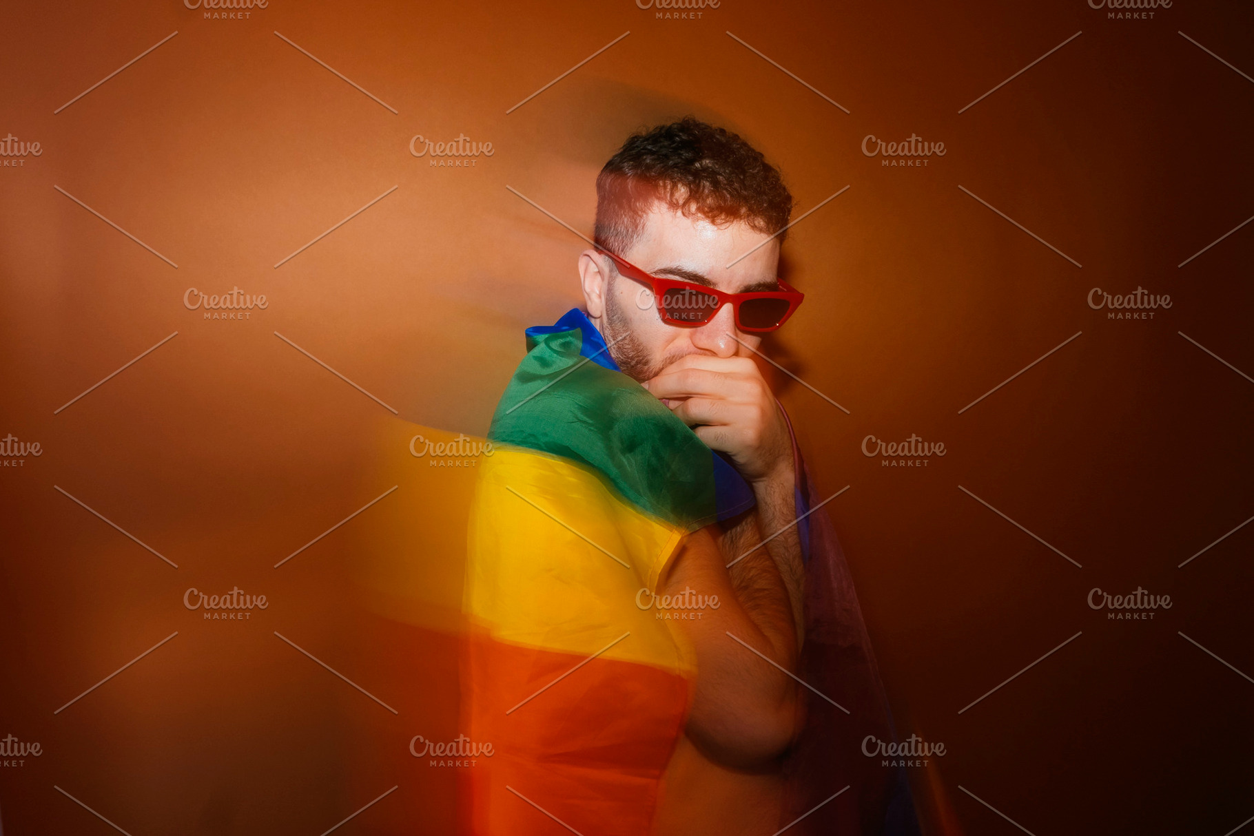 Young adult man wearing red sunglasses wrapped in rainbow flag, a Photo by karrastock