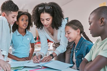 Female teacher in classroom with children, helping girl student with schoolwork and writing in book. Diversity in education, educator reading kids notebook and group learning together for assessment, a Photo by Peopleimages.com