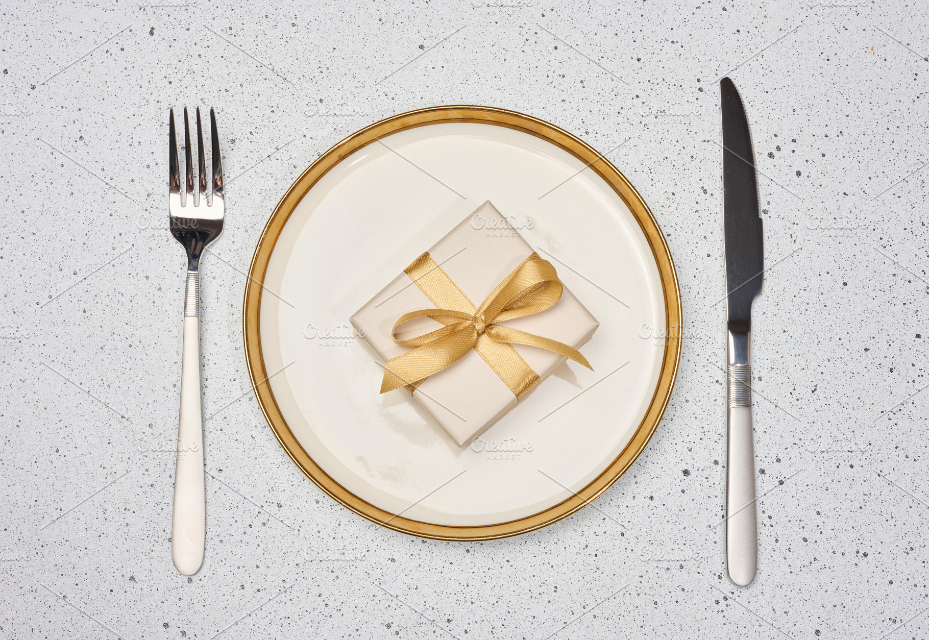 A gift with a gold bow on a white plate next to a fork and knife, a Holiday Photo by ndanko