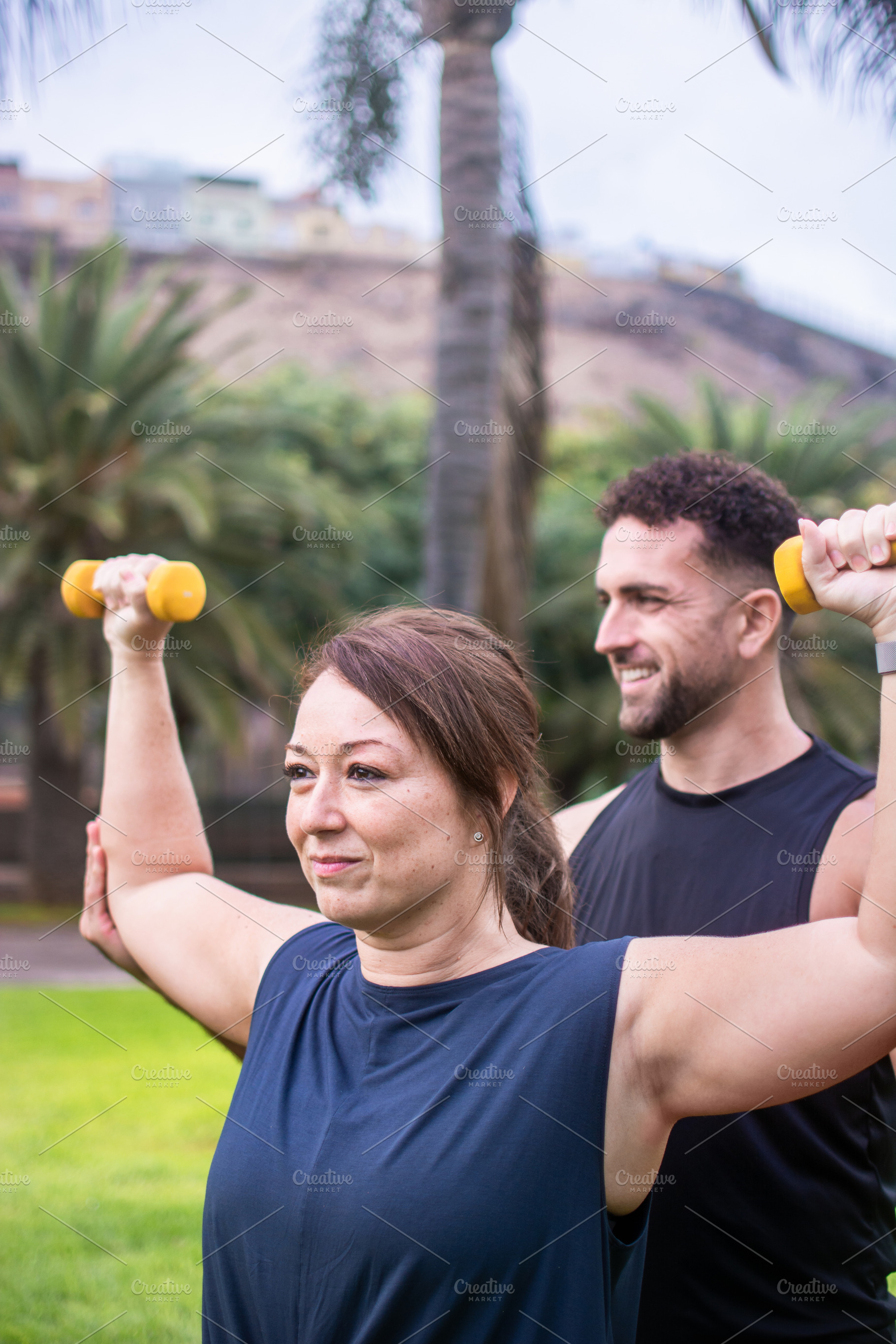 Male personal trainer assisting woman with dumbbell exercise outdoors, a Photo by etorres69