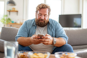 Overweight man enjoying snacks while using smartphone, a Person Photo by AlyaAnd