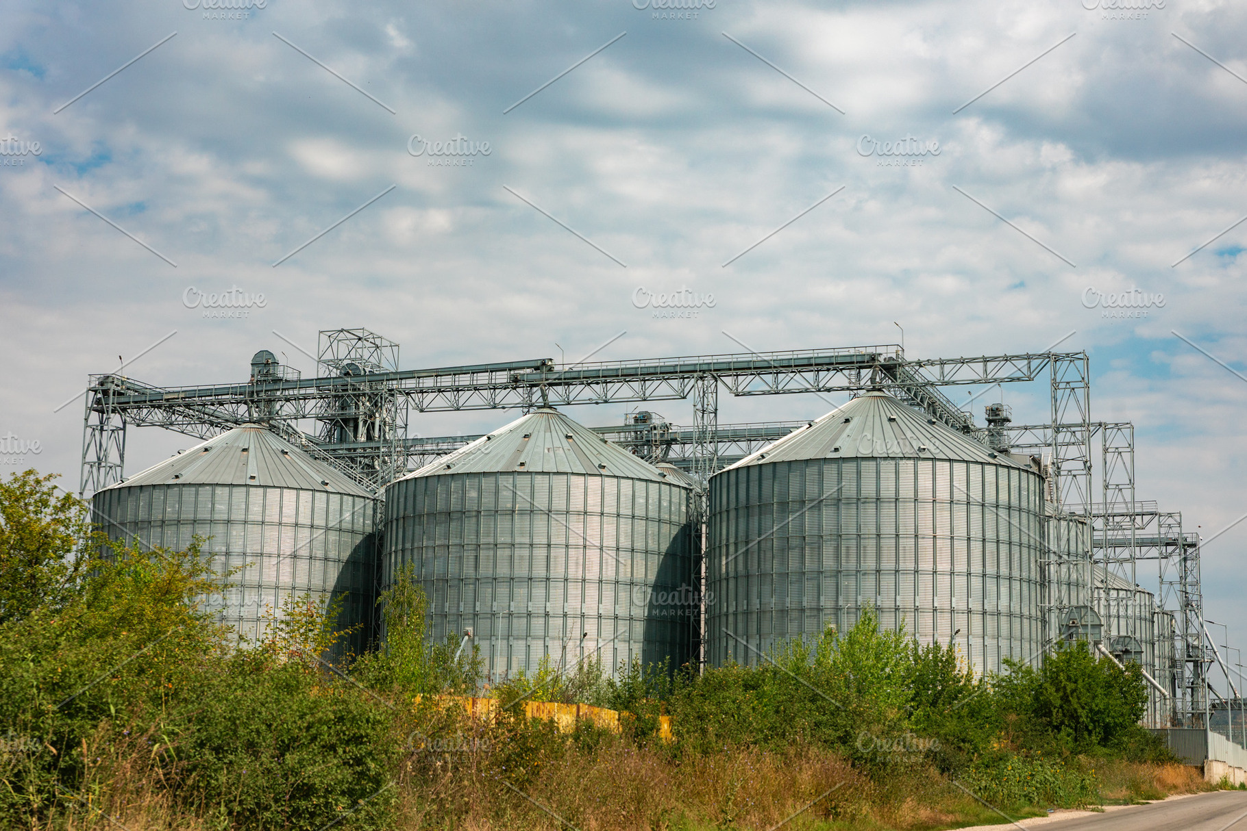 Large metal grain silos under a cloudy sky, surrounded by green vegetation, ill, an Industrial Photo by Ales Munt
