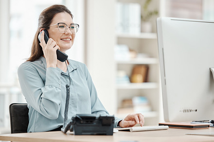 Telephone, computer and business woman in office, talking or conversation. Landline, receptionist and happy female from Canada in phone call, discussion or work call on desk in company workplace., a Photo by Peopleimages.com