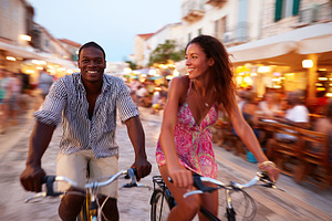 Couple enjoying an evening bike ride through a lively street in a coastal town, a Photo by AlyaAnd