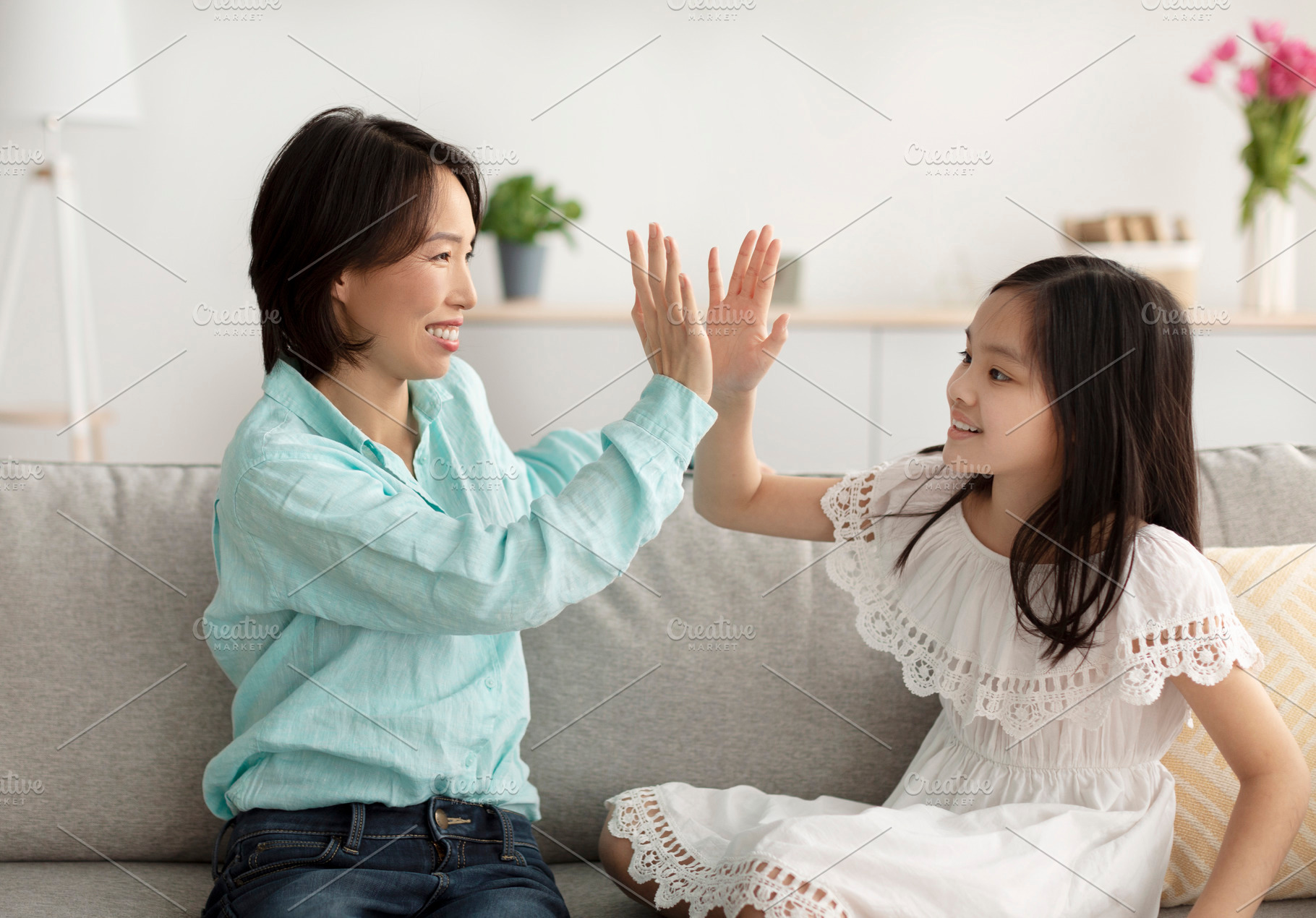 Cute little Asian girl giving high five to her grandmother, sitting on couch at, a Photo by Prostock-Studio