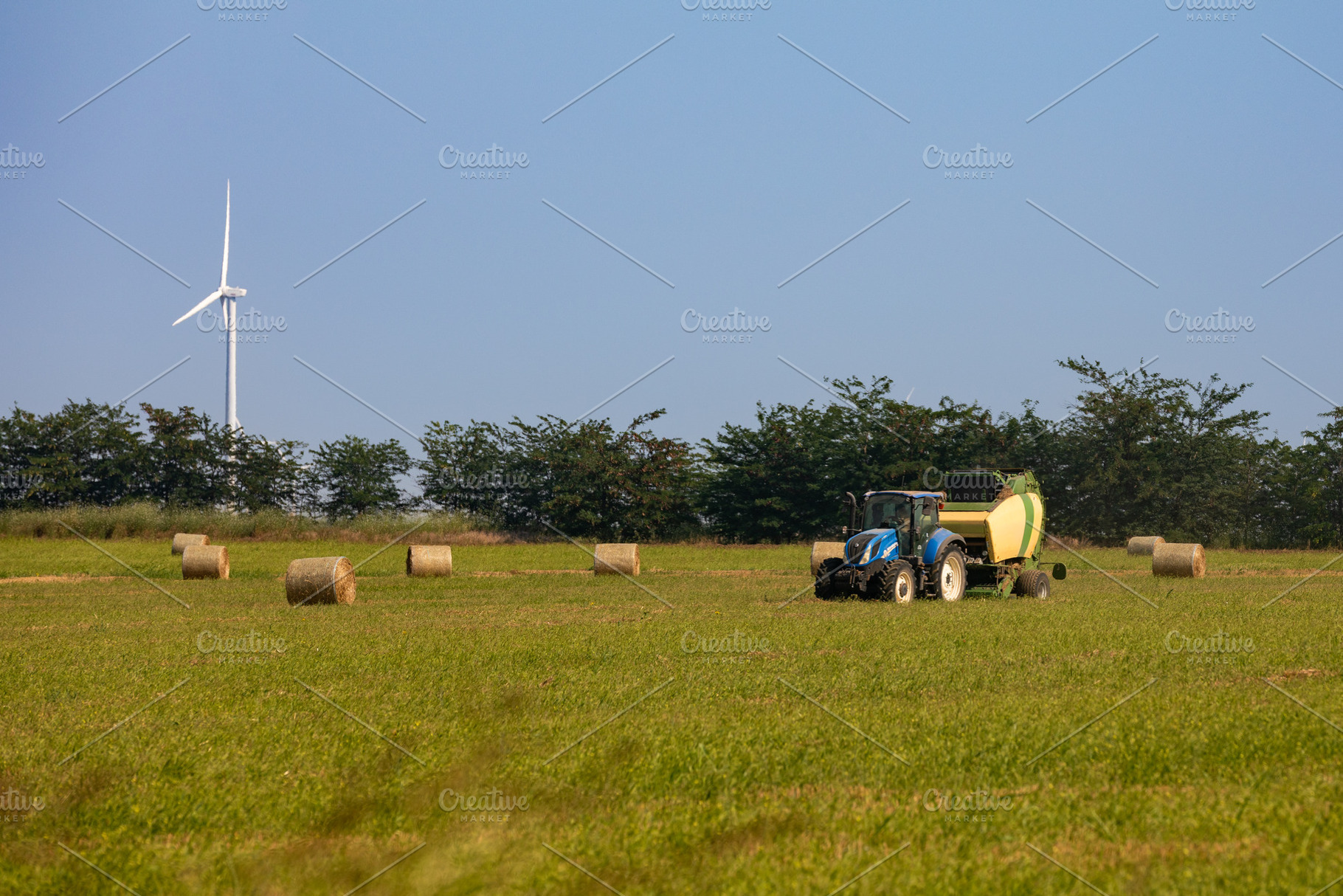 Modern tractor baling hay on a green field with round bales and a wind turbine, an Industrial Photo by Ales Munt