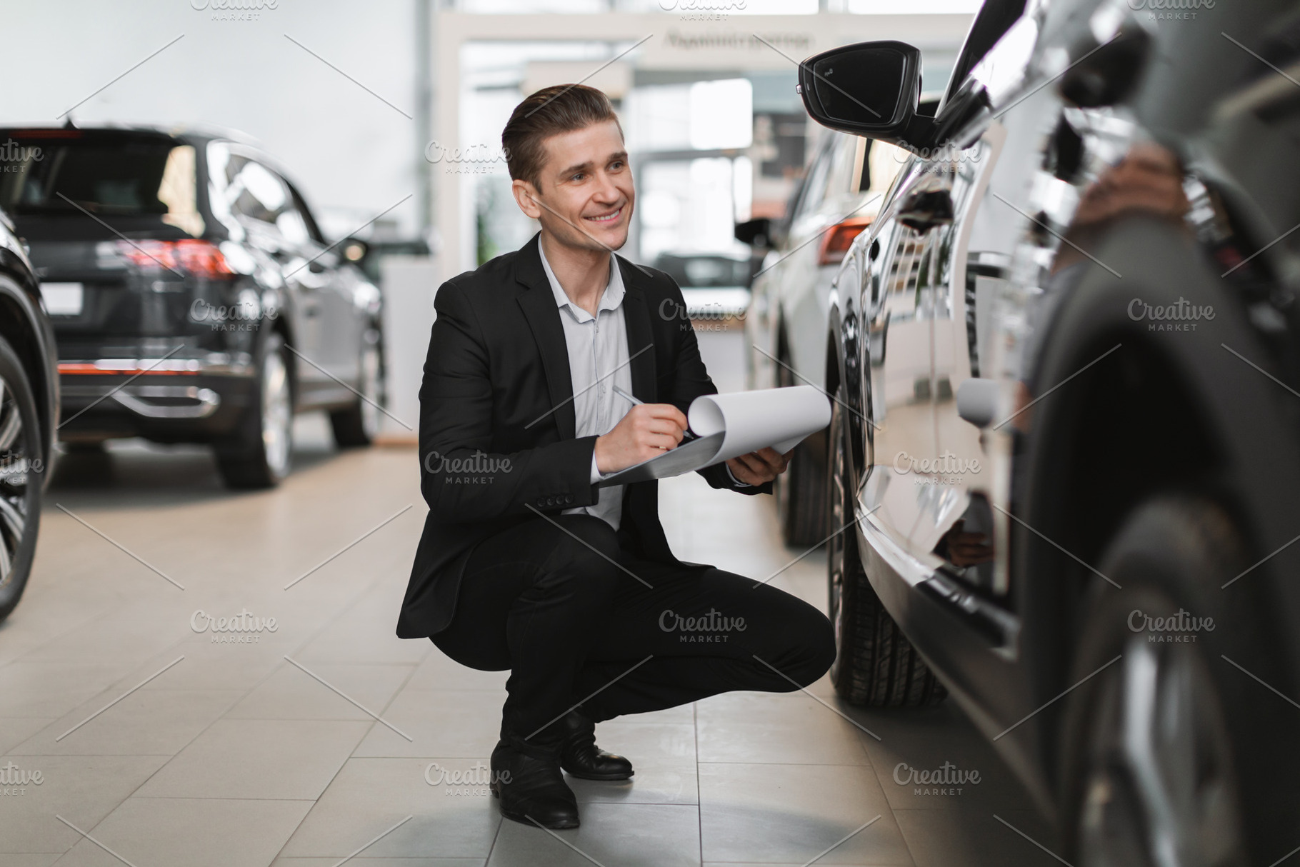 Happy young car salesman sitting near new auto, checking it, taking notes at au, a Person Photo by Prostock-Studio