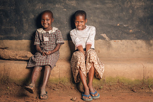 Two African Girls Sit & Smile at Hut