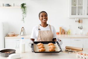 Satisfied young cute black woman in apron shows ready croissants in modern kitc, a Person Photo by Prostock-Studio