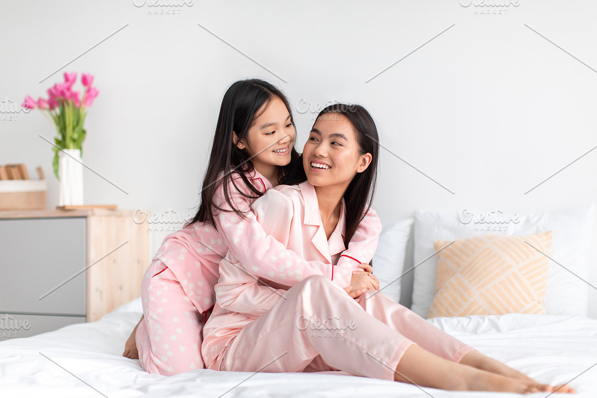 Smiling japanese teenage girl in pajama hugs young woman on bed and have fun in, a Photo by Prostock-Studio (Photo 2 of 2)