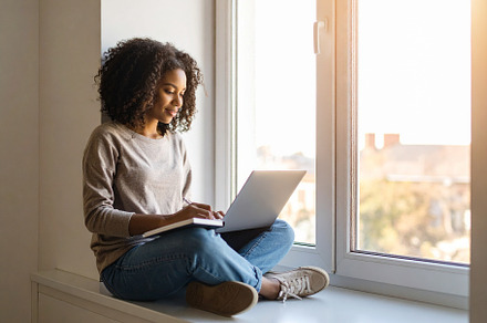 Student sitting by a window with a laptop and notebook, writing, a School & Education Photo by DanFLCreativo