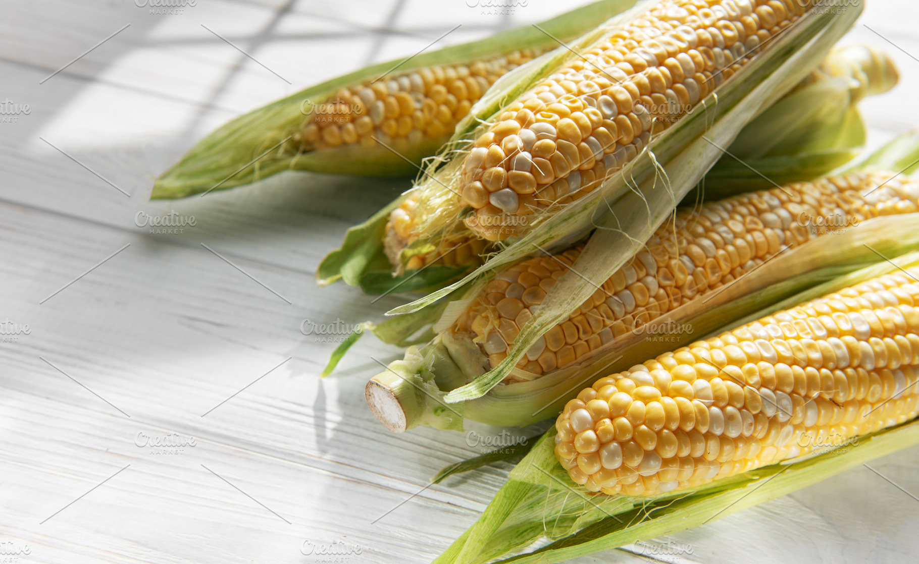 Fresh corn cobs with green husks on white wooden table, a Food & Drink Photo by Olena Rudo
