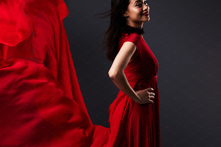Happy woman, art and fashion, red fabric on dark background with beauty and aesthetic movement. Flowing silk, fantasy and artistic model with smile in creative designer dress in studio with motion., a Photo by Peopleimages.com