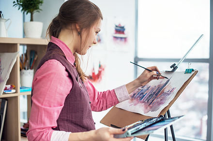 Side view portrait of a young woman painter drawing cityscape with watercolor palette on white paper using easel., an Arts & Entertainment Photo by undrey