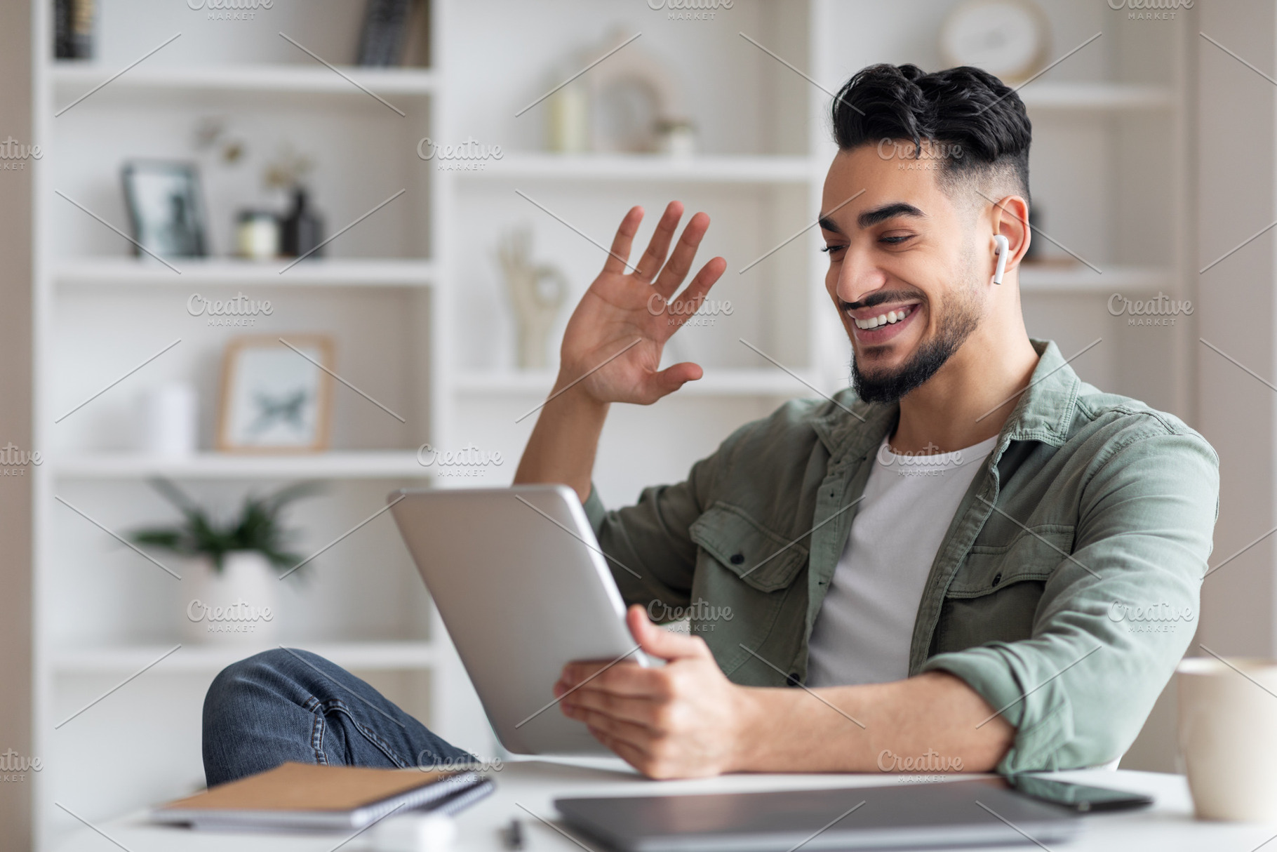 Happy young muslim guy with beard in wireless headphones looks at tablet and wa, a Photo by Prostock-Studio