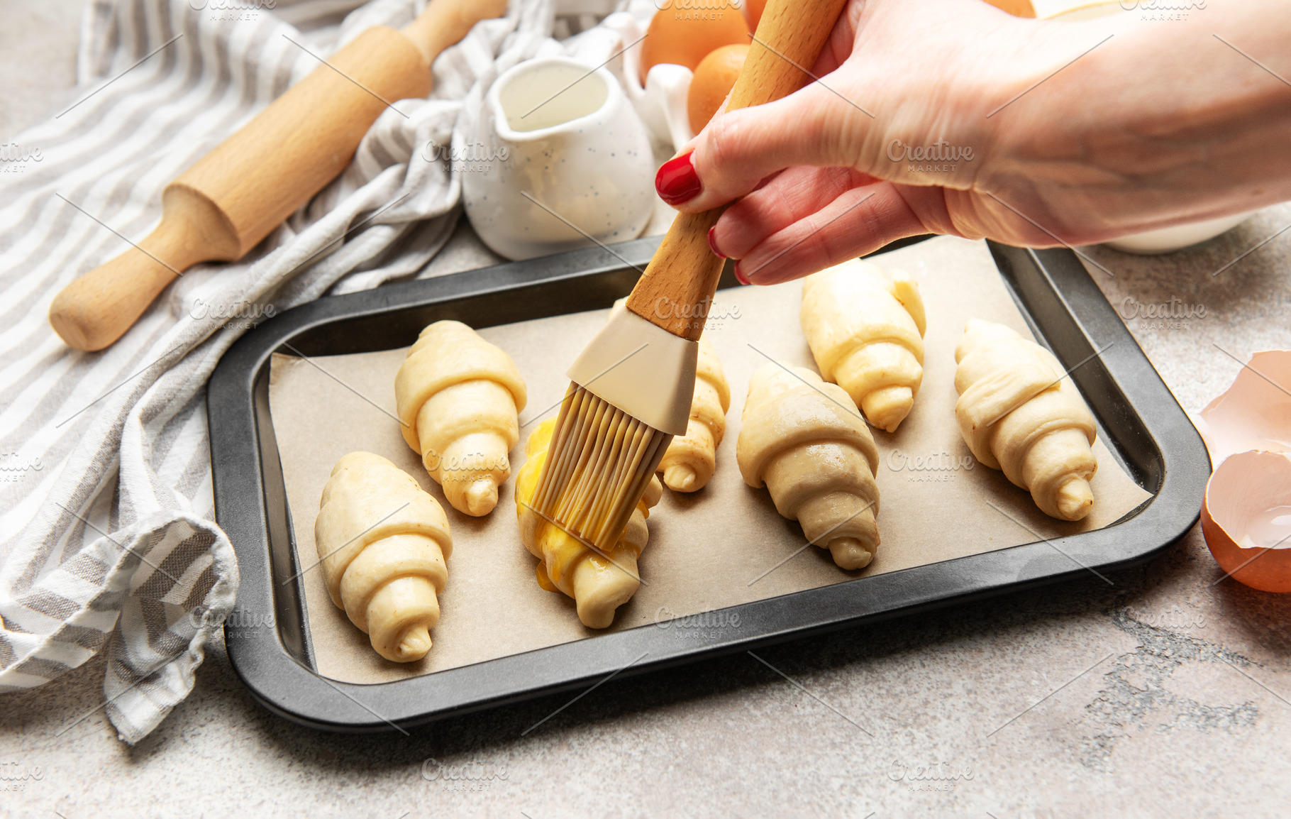 Baker brushing egg yolk on raw croissants on baking sheet, a Food & Drink Photo by Olena Rudo