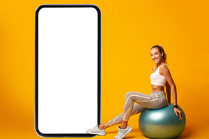 Sporty Lady Sitting On Fitball Near Phone Screen, Yellow Background, a Photo by Prostock-Studio