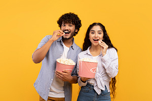 Excited indian man and woman eating popcorn at cinema, standing over yellow stu, a Person Photo by Prostock-Studio