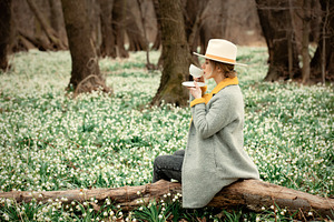stylish girl in gray coat with a cup in snowdrops meadow in fore