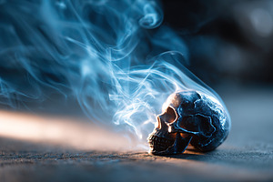 Dark smoke curls around a metallic skull on a dimly lit surface, a Photo by AlyaAnd