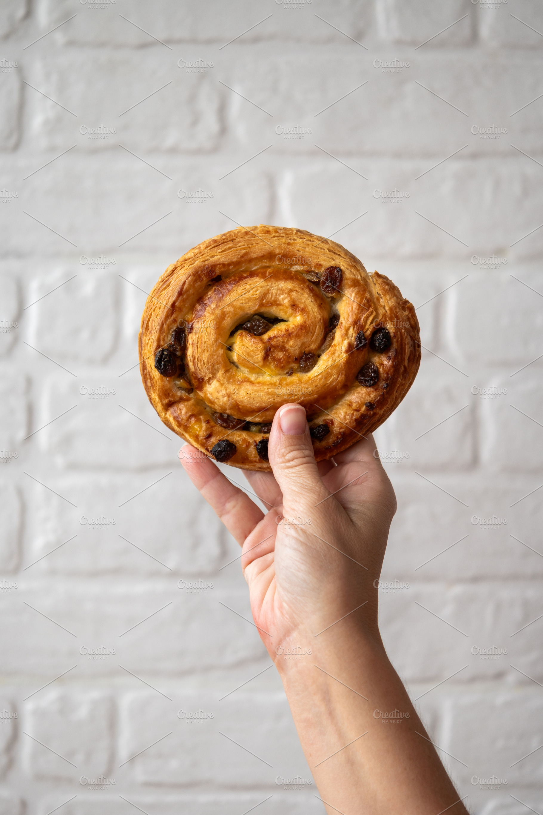 Hand holding pain aux raisins against white brick wall, a Food & Drink Photo by etorres69