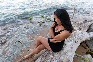 Smiling woman in black dress sitting on driftwood at rocky shoreline, enjoying, a Person Photo by Ales Munt