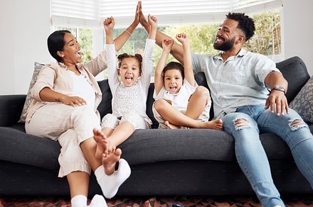 Happy parents and excited children celebrate on the sofa while watching television. Fun family time, relaxing at home and bonding. Mother and father high five, kids cheer for sports team win., a Photo by Peopleimages.com