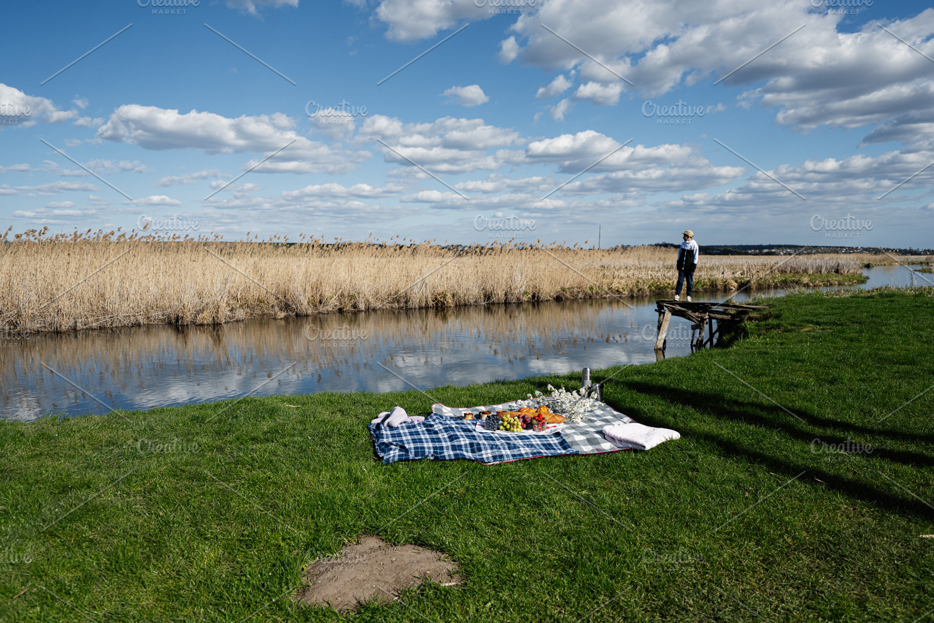 Boy standing on a rustic dock by a river with a picnic blanket laid out on gree, a Person Photo by AS photostudio
