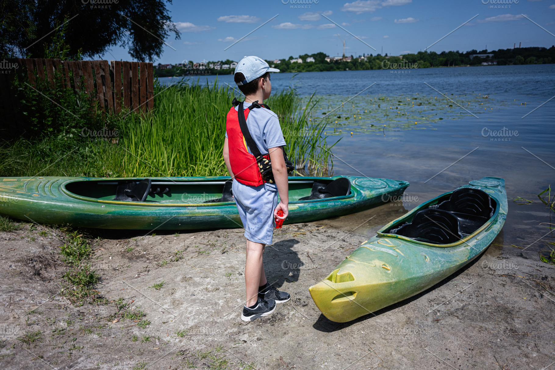 Boy standing on riverbank with kayaks ready for paddling under blue skies, a Photo by AS photostudio