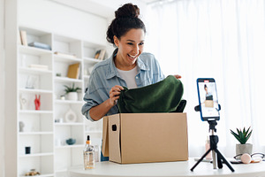 Female Blogger Making Video Unpacking Box With New Clothes Indoor, a Photo by Prostock-Studio