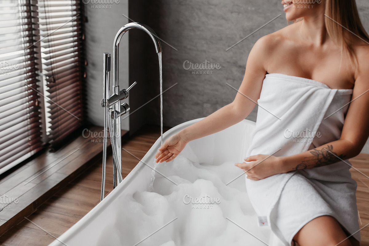 Young lady wrapped in towel preparing to take bath, sitting on bathtub and touc, a Photo by Prostock-Studio (Photo 4 of 5)