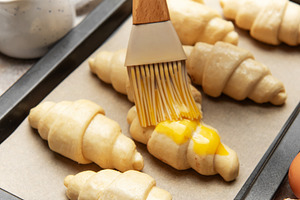 Baker brushing egg yolk on raw croissants on baking sheet, a Food & Drink Photo by Olena Rudo