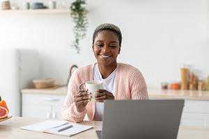 Happy millennial african american woman hold cup and work with laptop in modern, a Person Photo by Prostock-Studio
