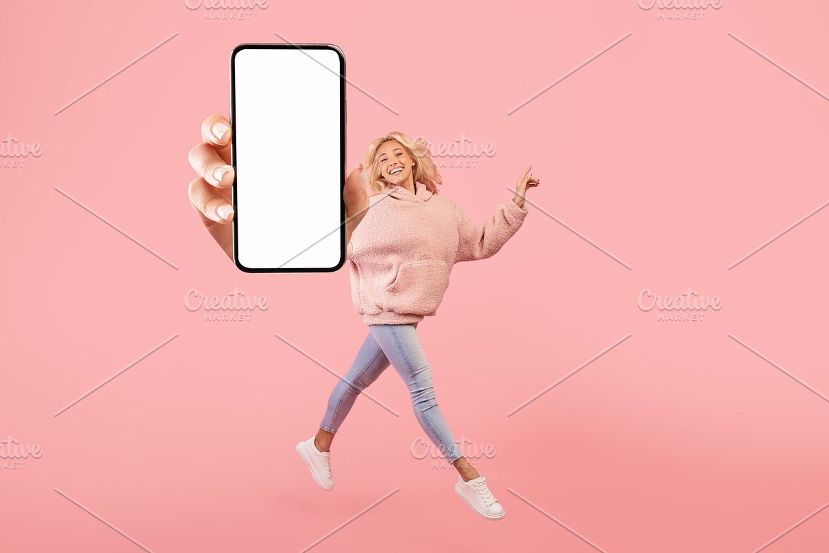 Happy young lady showing empty smartphone screen while jumping up over pink stu, a Photo by Prostock-Studio (Photo 1 of 3)