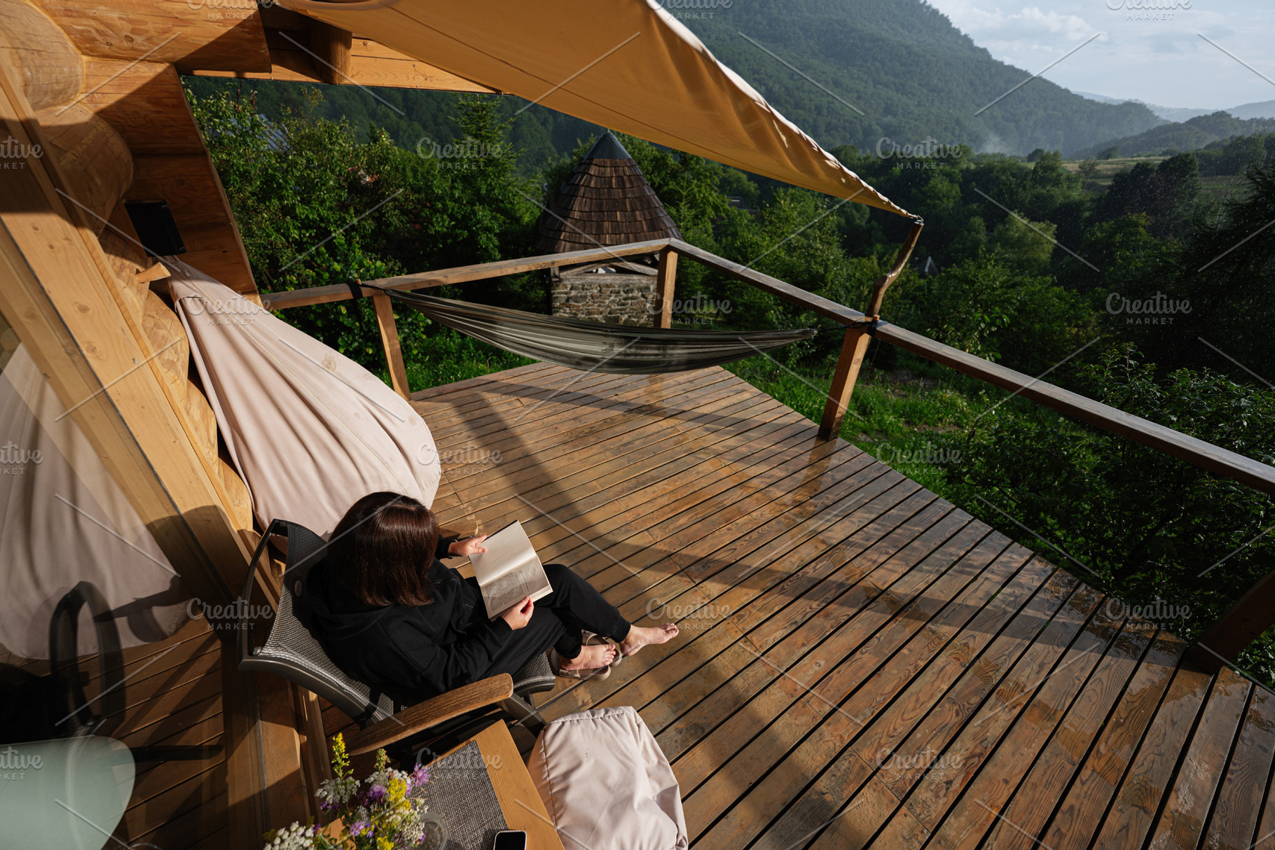 Woman reading a book on a wooden balcony overlooking a lush forest, a Photo by AS photostudio